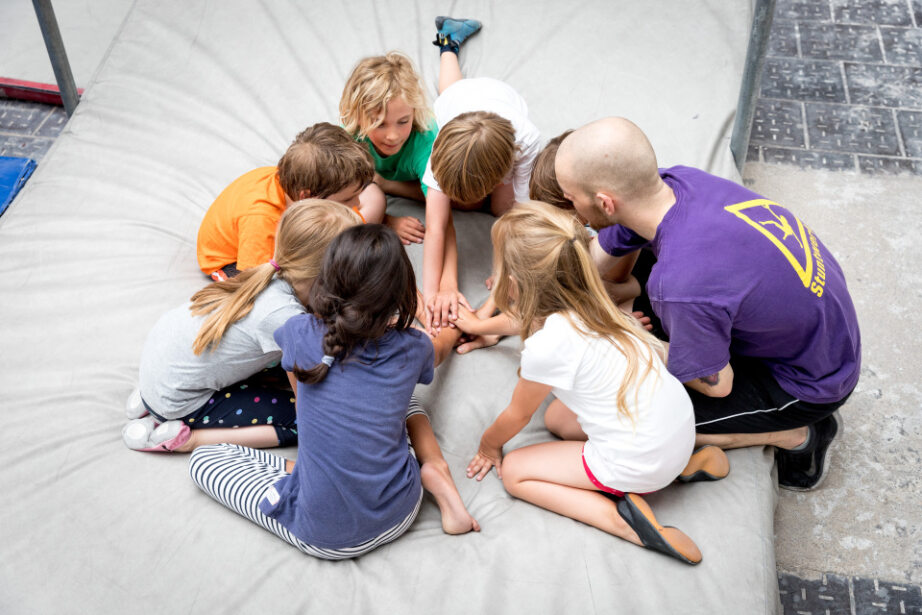 Kindergruppe im Stuntwerk Essen sitzt mit Trainer auf der Matte und legt gemeinsam die Hände zusammen. Spielerische Teambuilding-Situation im Kindertraining.